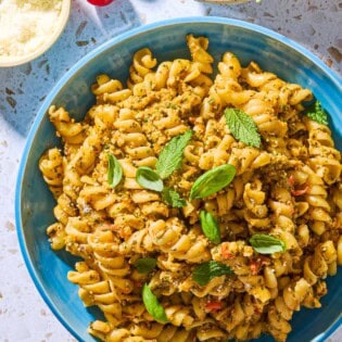 An overhead photo of pesto alla trapanese pasta in a bowl surrounded by 3 cherry tomatoes and bowls of pecorino romano cheese, and basil and mint.