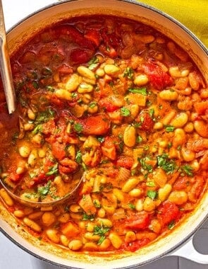 White bean stew in a large pot with a ladle next to a cloth napkin and a bowl of chopped parsley.