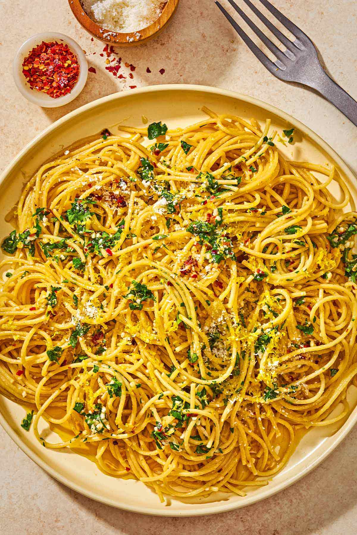 Lemon pasta topped with parsley, lemon zest, red pepper flakes and parmesan on a plate next to a fork and small bowls of red pepper flakes and parmesan cheese.