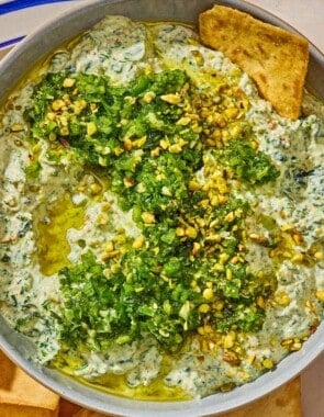 A bowl of spinach dip topped with pistachios and zhoug on a plate with pita chips.