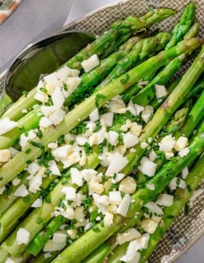Steamed asparagus topped with a vinaigrette, grated eggs and minced chives on a platter with a serving spoon. Next to this is a cloth napkin, a stack of 2 plates, and a bowl of minced chives.