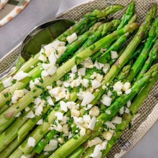 Steamed asparagus topped with a vinaigrette, grated eggs and minced chives on a platter with a serving spoon. Next to this is a cloth napkin, a stack of 2 plates, and a bowl of minced chives.