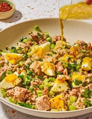 The dijon vinaigrette being poured onto the tuna salad with egg in a serving bowl with a small bowl of aleppo pepper in the background.