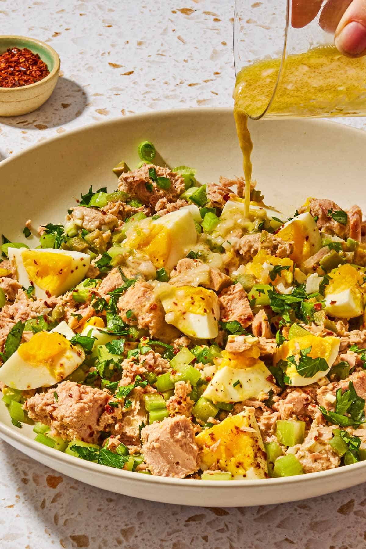 The dijon vinaigrette being poured onto the tuna salad with egg in a serving bowl with a small bowl of aleppo pepper in the background.
